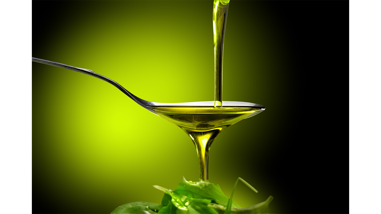 A spoonful of olive oil being poured over fresh green leaves against a dark background.